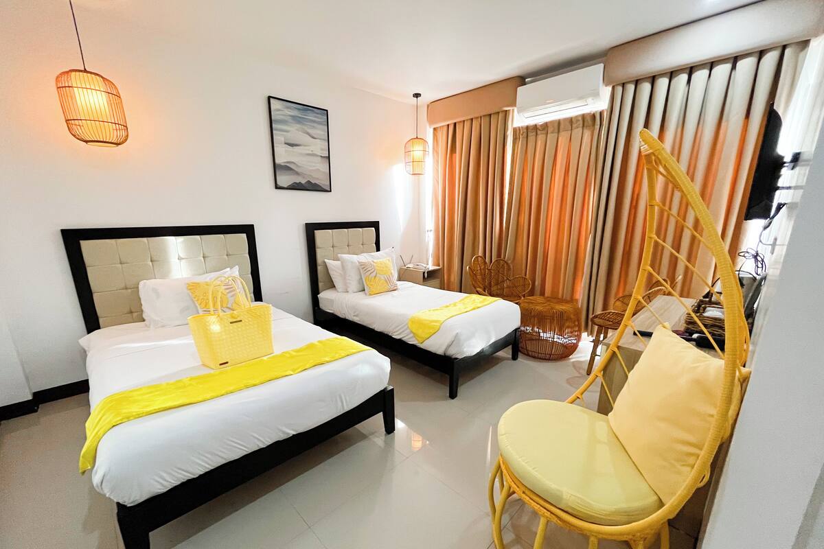 A hotel room with two beds, a chair, a hanging light, and a window with curtains.