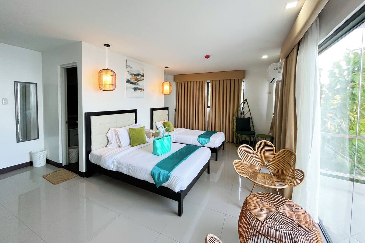 A hotel room with two beds, a sitting area with wicker furniture, and a view of greenery through the window.