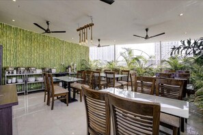 Restaurante con decoración de pared de bambú, varias mesas y sillas, ventiladores de techo y grandes ventanales con vistas a un paisaje urbano.