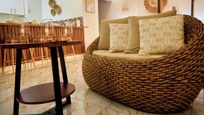 Un sofá de mimbre con cojines estampados con motivos de palmera, una mesa de comedor de madera con sillas y una zona de cocina con armarios y una lámpara colgante.