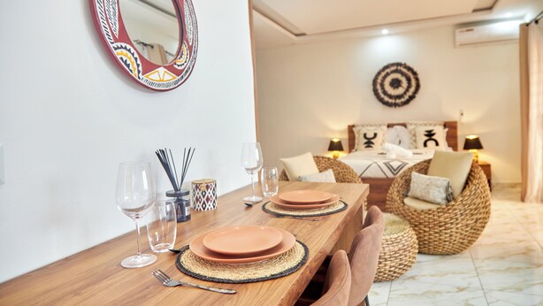 Un comedor con una mesa de madera dispuesta para dos, un espejo decorativo y una cama con cabecera al fondo.