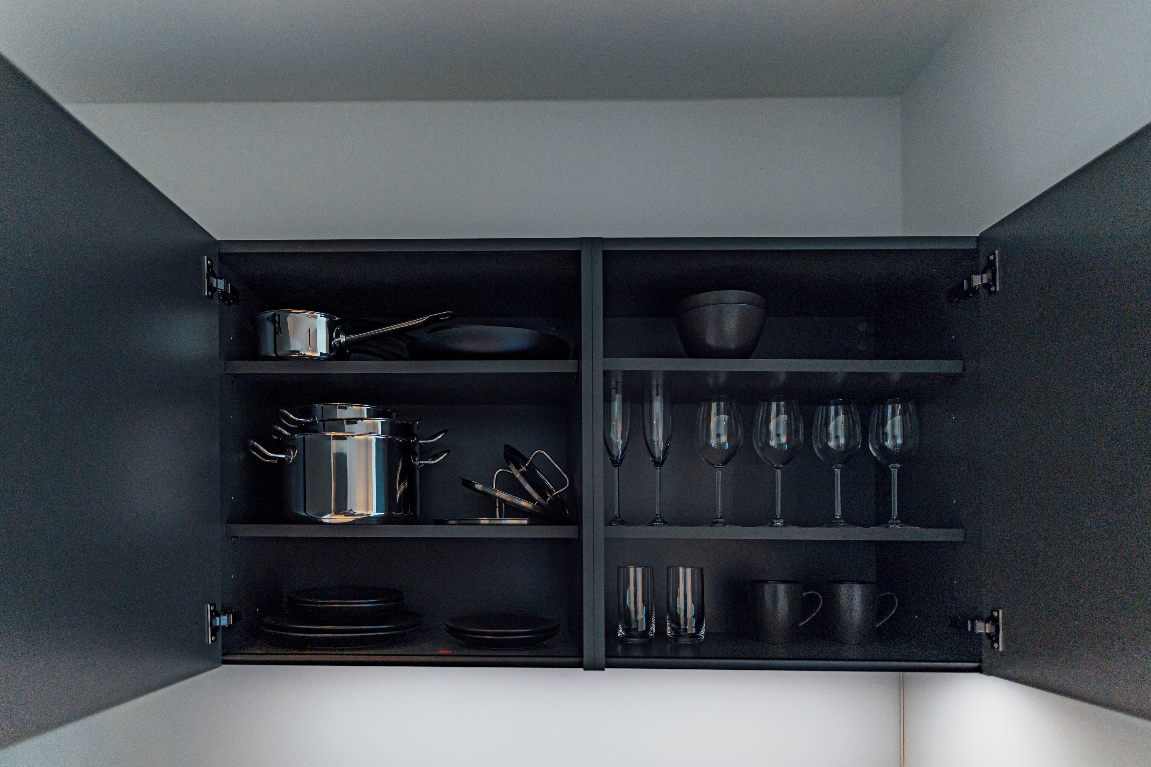 A kitchen cabinet with shelves containing cookware, plates, bowls, wine glasses, and cups.