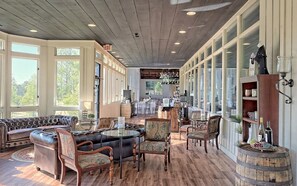 A spacious living area with a leather sofa, wooden chairs, and a dining table. There are large windows providing natural light and a view of the outdoors.