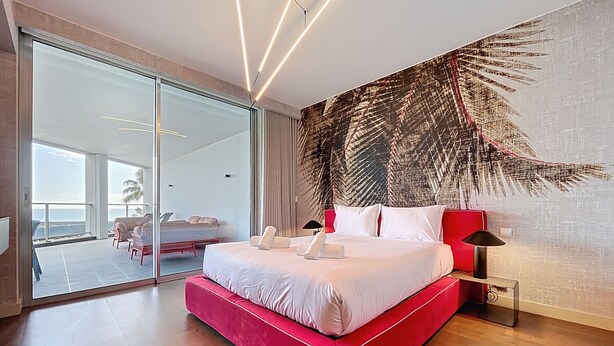 Un dormitorio moderno con una cama grande, una puerta de vidrio que da a un balcón y un mural de hojas tropicales en la pared.