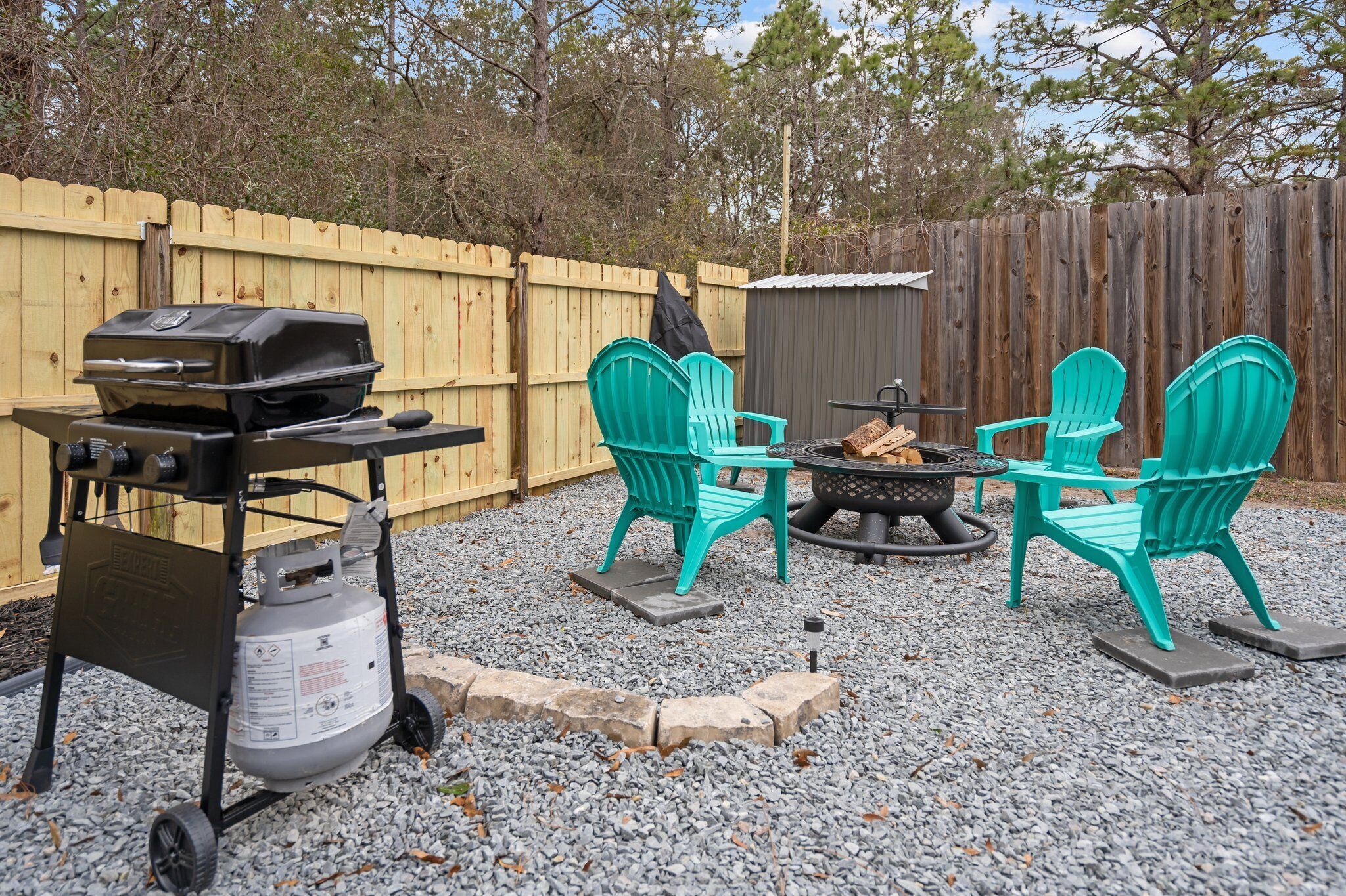 Un jardin avec un barbecue, deux chaises vertes, un foyer et une clôture en bois.