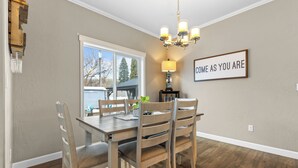 A dining area with a wooden table and chairs, a chandelier, and a window with a view of trees and a building.