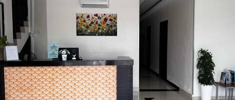 A reception area with a patterned reception desk, an air conditioning unit, a floral painting, and a potted plant.