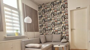 Una sala moderna con un sofá seccional gris, una mesita redonda pequeña, un tapete gris, un radiador, una ventana con persianas, y una pared con papel tapiz con un motivo urbano.