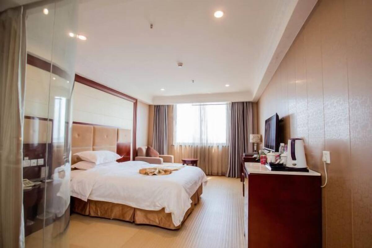 A hotel room with a large bed, a desk with a TV, and a glass partition.