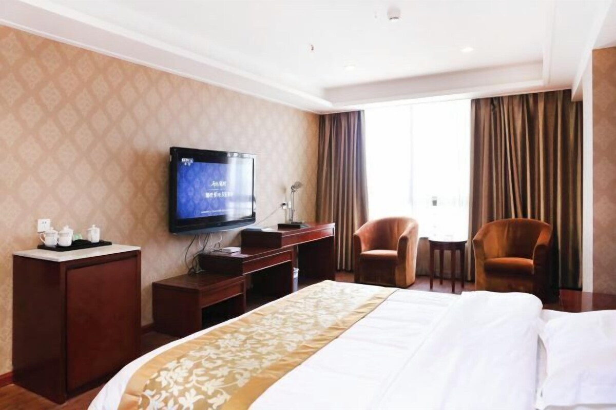 Une chambre d’hôtel avec un grand lit, un téléviseur fixé au mur, un bureau avec une chaise et une vue sur l’extérieur par une fenêtre.
