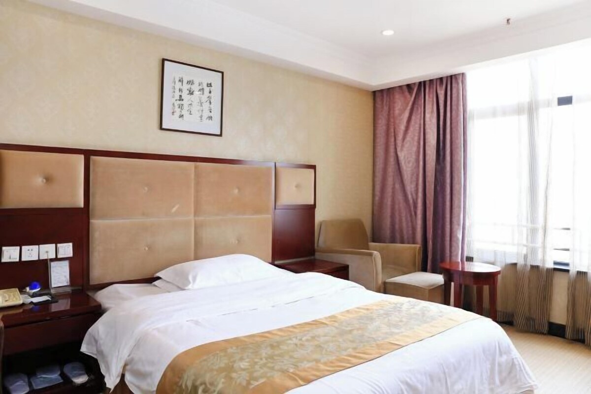 Hotelkamer met een groot bed, nachtkastje, stoel, een klein tafeltje en een raam met gordijnen.