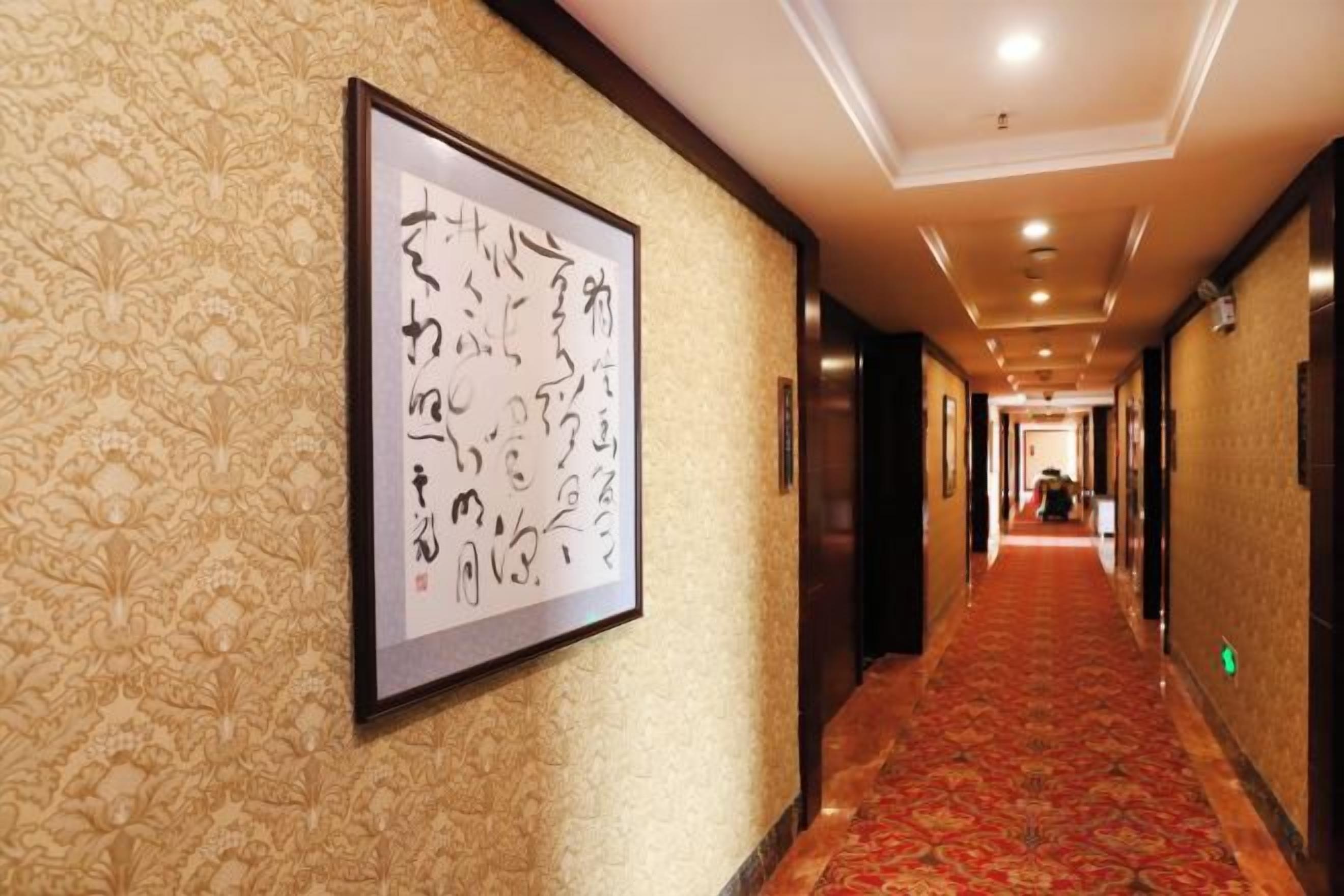 A hotel corridor with patterned wallpaper, a framed artwork on the wall, and a red carpet.