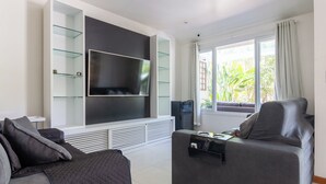 Ein modernes Wohnzimmer mit großem Flachbildfernseher, integrierten Regalen, einem grauen Sofa und einem Fenster mit Vorhängen.