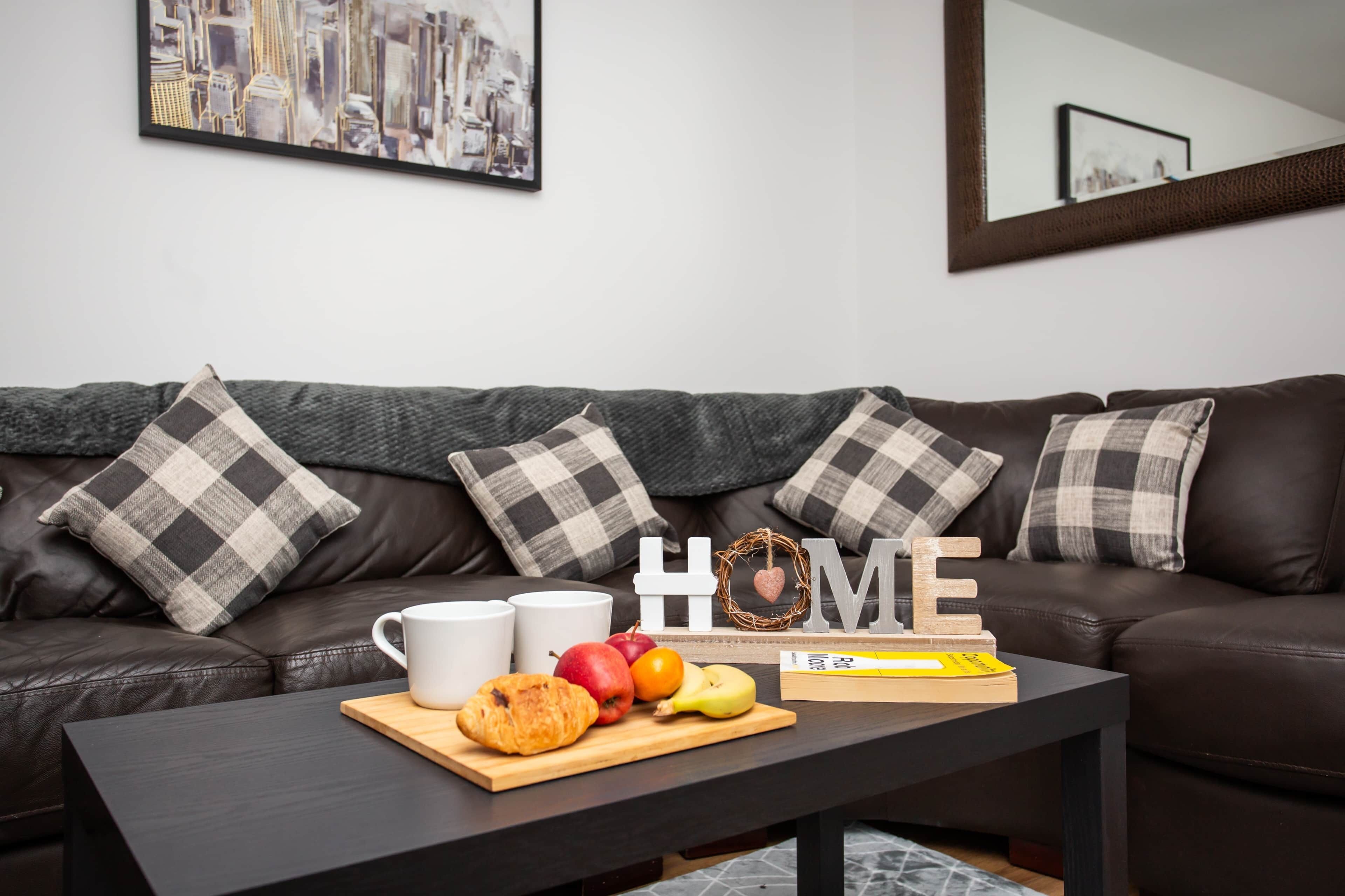 A dark leather sofa with checkered pillows, a wooden coffee table with food and a book, and a framed cityscape painting on the wall.