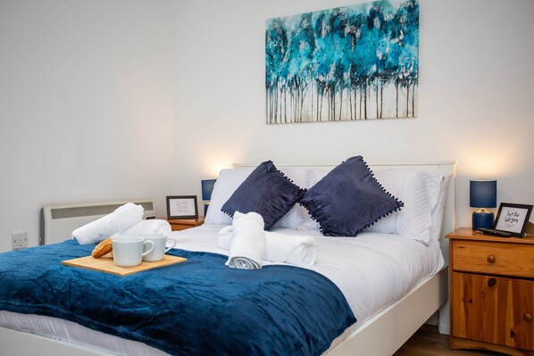 A neatly made bed with a blue blanket, white pillows, and a wooden tray with a mug and croissant. There is a painting on the wall and a nightstand with a lamp and a framed picture.