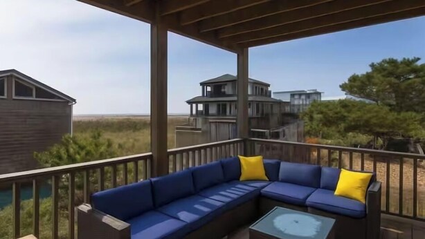 A covered outdoor deck with a blue sectional sofa and a glass-top coffee table. - Gatsby’s Main Level. Ocean Bay Park. 
Heated Saltwater Pool + Hot Tub. Sleeps 14 (Ocean Beach)