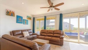 A spacious living room with a brown leather sofa, a coffee table, and a ceiling fan. There are large windows with curtains and a view of a house outside. - Gatsby’s Main Level. Ocean Bay Park. 
Heated Saltwater Pool + Hot Tub. Sleeps 14 (Ocean Beach)