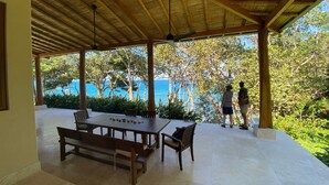 Une terrasse extérieure couverte, dotée d’une table en bois et de bancs, offrant une vue panoramique sur la mer et une végétation luxuriante.