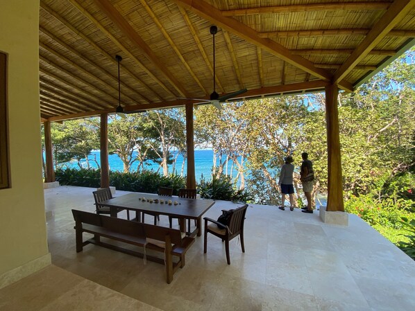 An outdoor covered patio with a wooden table and benches, overlooking a scenic view of the sea and lush greenery.