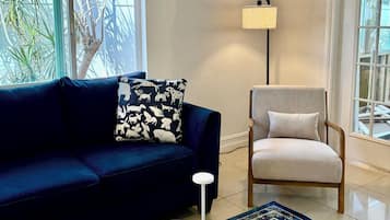 Un salón con un sofá azul oscuro, un sillón de color claro, una mesa de centro con un control remoto y revistas, una alfombra estampada y una lámpara de pie.