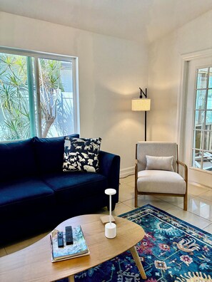 A living room with a dark blue sofa, a light-colored armchair, a coffee table with a remote control and magazines, a patterned rug, and a floor lamp.