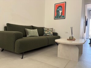 Un canapé vert avec deux coussins, une table ronde blanche ornée d’un objet décoratif et une œuvre d’art encadrée accrochée au mur.