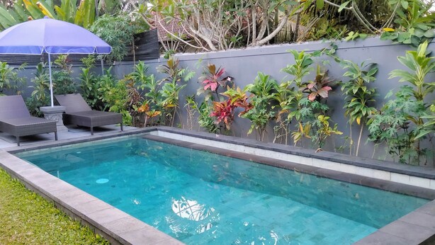 A rectangular swimming pool with a clear blue water surface, surrounded by a low stone wall. There are two dark-colored lounge chairs and a blue umbrella providing shade. - Umah Landuh, Spacious and comfortable private house near Ubud city (Bali)