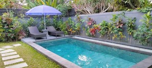 Une piscine rectangulaire à l’eau d’un bleu limpide, entourée d’un petit muret en pierre. On y trouve deux chaises longues de couleur sombre et un parasol bleu qui offre de l’ombre.