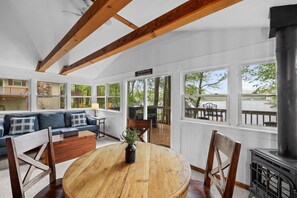 A spacious living room with a wooden dining table, a sofa, and a fireplace. Large windows offer views of the outdoors. - Lakefront Retreat | Deck, AC, Fire Pit, BBQ, Wi-Fi (Pocono Lake)