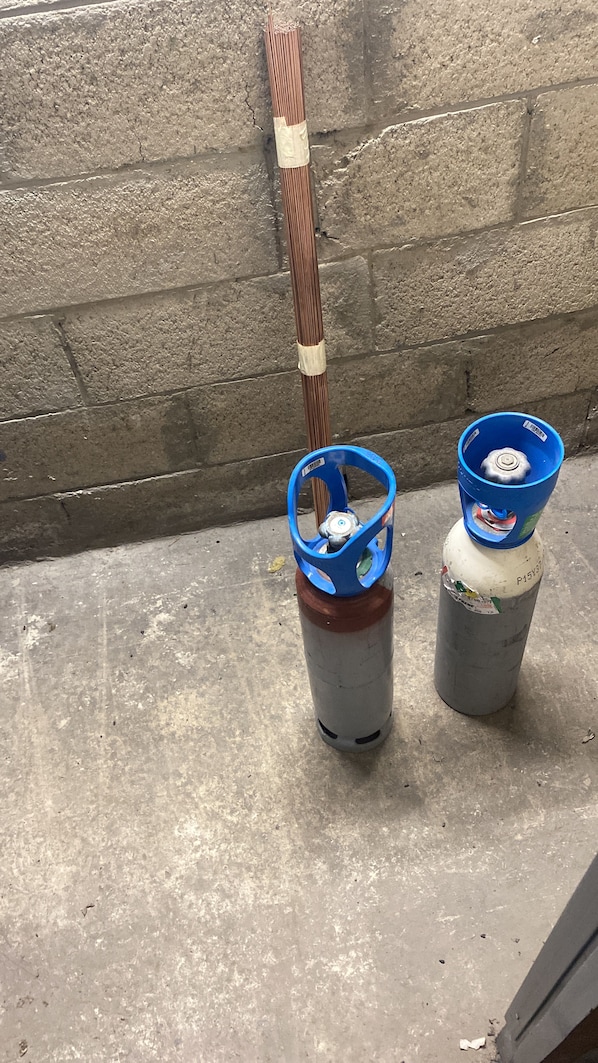 Two gas cylinders with blue tops, one with a white label and the other with a blue label, placed on a concrete floor against a brick wall.