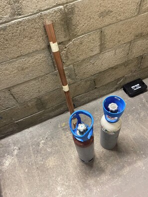 Two gas cylinders with blue caps and a metal pipe against a concrete wall.