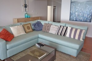 Un sofá seccional azul claro con varios cojines estampados, una mesa de centro de vidrio con un libro y una mesita de noche de madera con botellas decorativas.