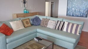 A light blue sectional sofa with various patterned cushions, a glass coffee table with a book, and a wooden side table with decorative bottles. - Large waterfront home with bay views to Moreton Island (Amity Point)