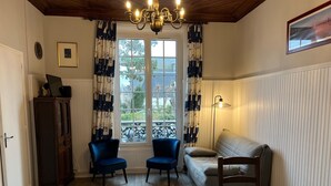 A room with a wooden floor, a sofa, two chairs, a table, a cabinet, a chandelier, a window with curtains, and a picture on the wall. - Ground floor apartment in old villa 60 m from the beach (Cabourg)