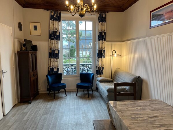 Une pièce avec un plancher en bois, un canapé, deux fauteuils, une table, un buffet, un lustre, une fenêtre avec des rideaux et un tableau au mur.