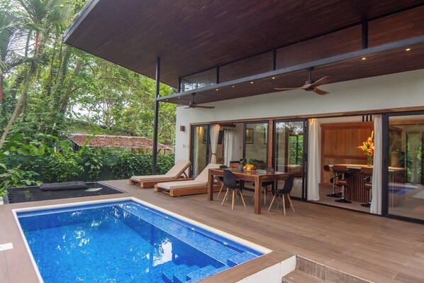 A modern outdoor pool area with a wooden deck, lounge chairs, and a dining table.