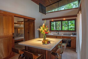 A modern kitchen with a central island, wooden cabinets, a large window with a view of greenery, and a dining area with chairs.