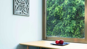 A corner desk with a view of a forest through a window, a decorative metal piece on the wall, and a plate with apples on the desk. - Raphael City Nest (Tel Aviv)