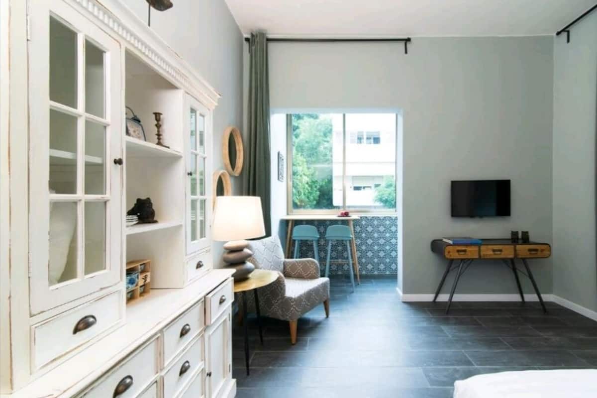 A hotel room with a large white cabinet, a small round table with a lamp, a sofa, a TV on a stand, and a window with a view of greenery.