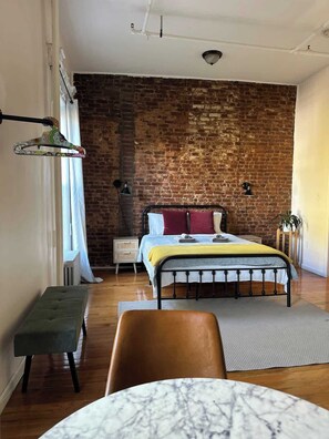 Une chambre avec un mur en briques, un lit avec une tête de lit, des tables de chevet, un banc, une chaise et une table en marbre.