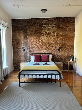 A bedroom with a brick wall, a bed with a metal frame, two bedside tables, and a floor lamp.. Brooklyn lovely studio apartment!