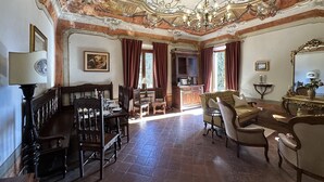 A room with a dining table, chairs, a sofa, and a desk. There is a chandelier on the ceiling.