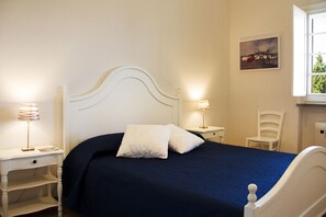 A bedroom with a white bed, two bedside lamps, a nightstand, a chair, and a painting on the wall. - Le Duchesse Rooms and Events (Trani)