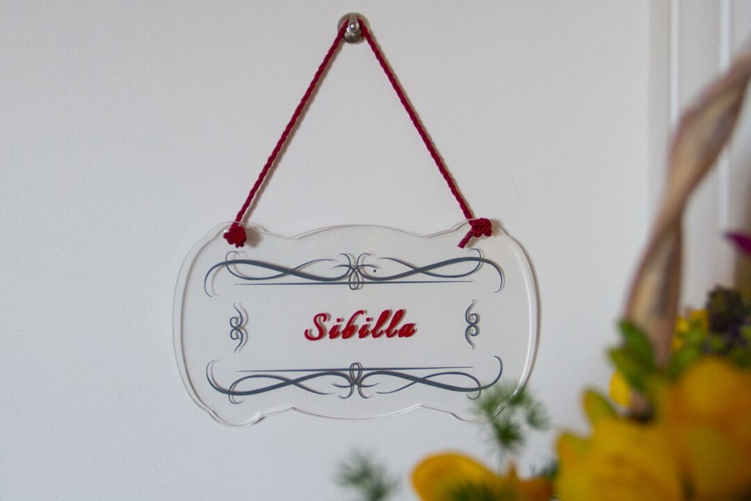 A clear glass sign with the name "Sibilla" in red lettering, hanging from a red string.