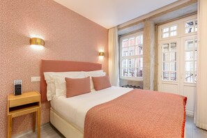 A hotel room with a bed, a nightstand, a wall-mounted light, and a window with a view of buildings. - Charming Riverview Apartment in Porto (Porto)