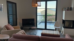 A living room with a pink sofa, a television, a coffee table, and a fireplace. There is a large window with a view of a pool and a building. - Air-conditioned villa with pool and panoramic sea view, 6 pax (Brando)