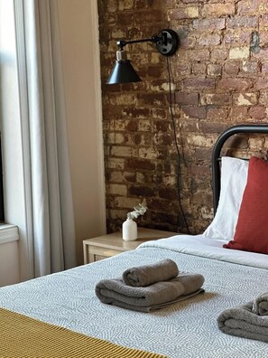 Habitación de hotel con pared de ladrillo, cama con almohadas blancas y rojas, mesita de noche con un jarrón y una lámpara.