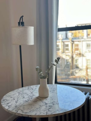 Una mesa redonda de mármol con un jarrón blanco que contiene follaje verde, ubicada junto a una ventana con vistas a edificios.