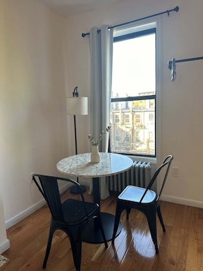 Una habitación con una mesa redonda, dos sillas, una lámpara de pie, un jarrón con flores, una ventana con cortinas y un radiador.