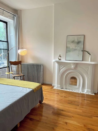 A bedroom with a bed, a radiator, a fireplace, and a window.. Brooklyn shoebox studio apartment!